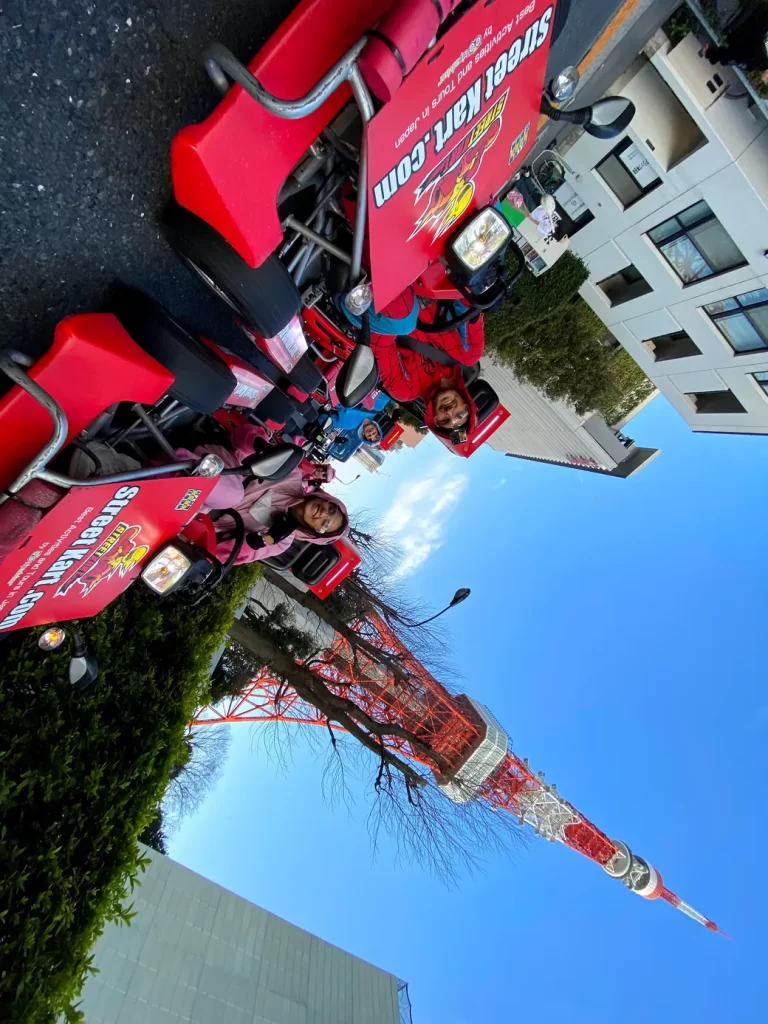 People in red outfits riding red promotional go-karts with banners along a street; Tokyo Tower visible in the background.