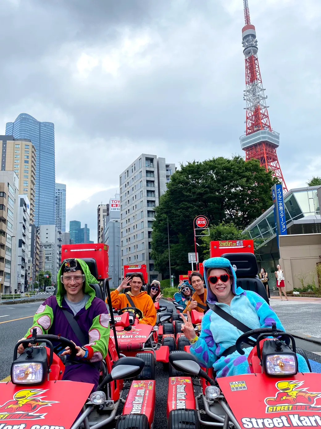 Group of friends in bright go-karts racing down a city street with Tokyo Skytree in the background, smiling at the camera on a cloudy day.
